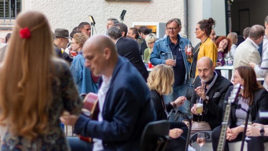 Viele stehende oder sitzende Leute unterhalten sich. Von hinten ist die Band zu sehen, wie sie performt.